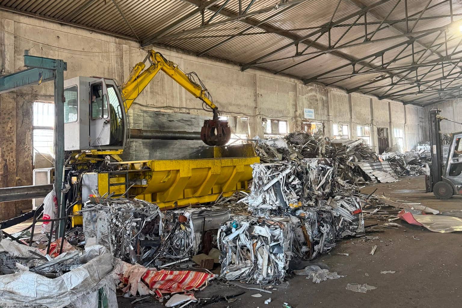 Grajfer sa žutom rukom iznad prese, uz složene bale aluminija i viljuškar u dnu hale