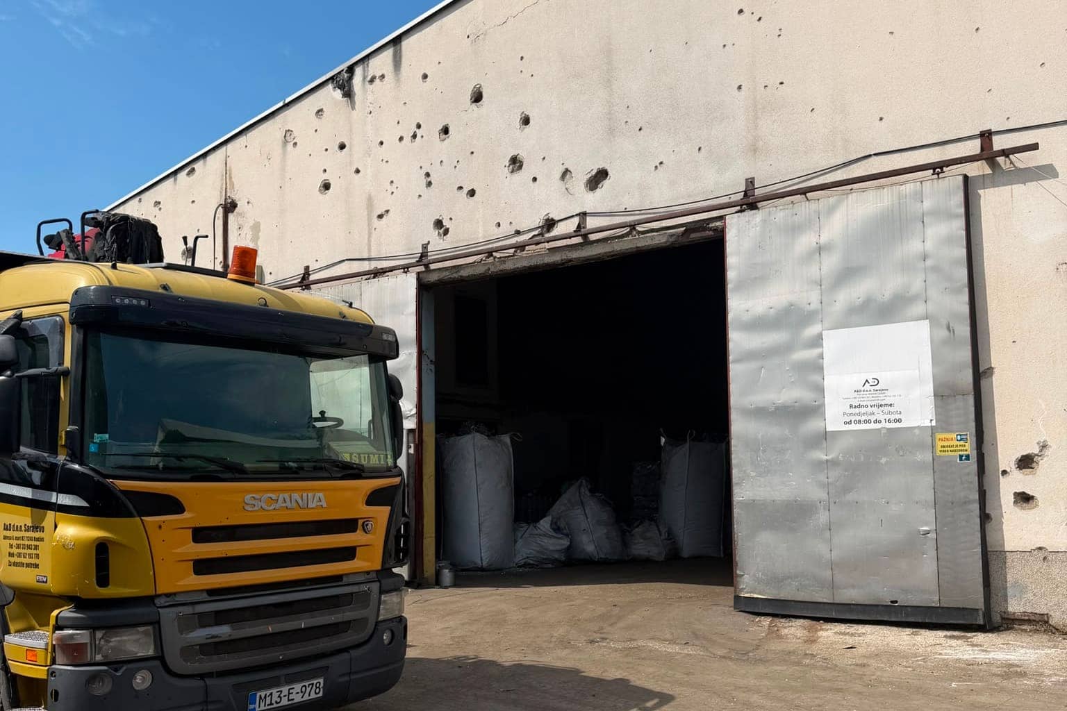A yellow truck parked at the open hall door, which carries a sign with the opening hours